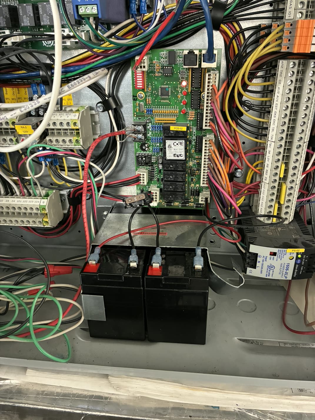 CaptiveAire control cabinet interior — the green CORE PCB control board (Rev 1.63) mounted above two black 12V 7Ah sealed lead-acid batteries (PS-1270-F2) wired in series with red and black faston connectors, surrounded by terminal strips, relay modules, an Allen-Bradley 1606-XLP 24V DC power supply, and rainbow-colored field wiring